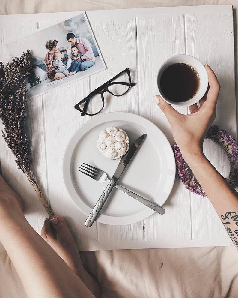 May include: A white plate with a fork and knife, a small white cake with a brown topping, and a cup of coffee on a white wooden surface. A person's arm with a tattoo is visible in the background.