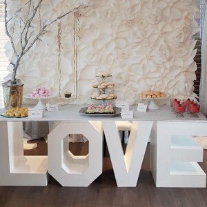 Könnte beinhalten: Weiße LOVE-Buchstaben, die das Wort "LOVE" buchstabieren, werden als Desserttisch für eine Hochzeit oder Party verwendet. Der Tisch ist mit weißen Blumen, Cupcakes und anderen Leckereien dekoriert.