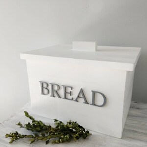 Puede incluir: Caja de pan de madera blanca con una etiqueta "BREAD" gris en la parte delantera. La caja tiene una tapa con bisagras y est&aacute; sobre una superficie de madera blanca.