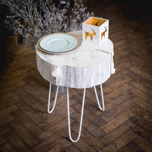 May include: White wooden side table with a natural wood grain finish. The table has a round top and three hairpin legs. A white ceramic candle holder with a reindeer design sits on the table, along with a light blue plate.