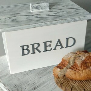 Puede incluir: Caja de pan de madera blanca con tapa gris y la palabra "BREAD" en letras grises en la parte delantera. Una hogaza de pan est&aacute; sentada sobre una tabla de cortar de madera delante de la caja.