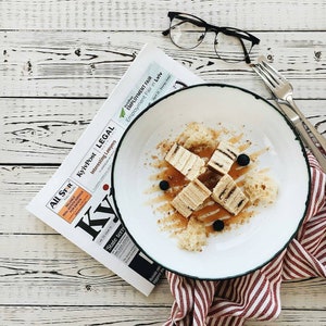 May include: A white plate with a green rim contains a dessert of layered cake squares, drizzled with honey and topped with blueberries. The plate is on a red and white striped napkin, next to a newspaper and a pair of glasses.