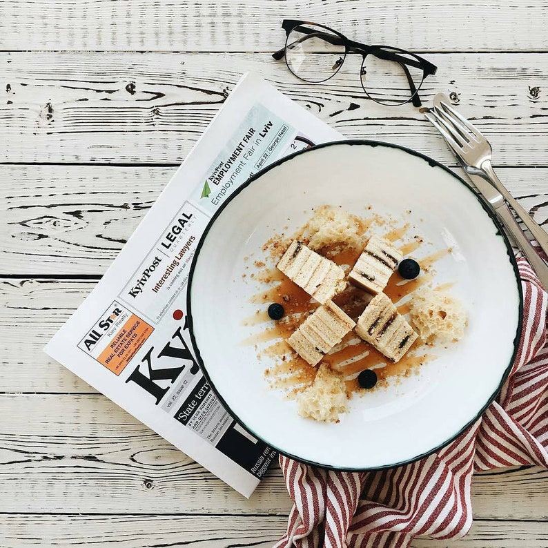 May include: A white plate with a green rim contains a dessert of layered cake squares, drizzled with honey and topped with blueberries. The plate is on a red and white striped napkin, next to a newspaper and a pair of glasses.