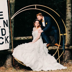 May include: A bride and groom pose for a wedding photo in a forest setting. The bride is wearing a white wedding dress and the groom is wearing a dark suit. They are standing in front of a gold metal archway decorated with greenery. The sign behind them reads "To the moon and back".