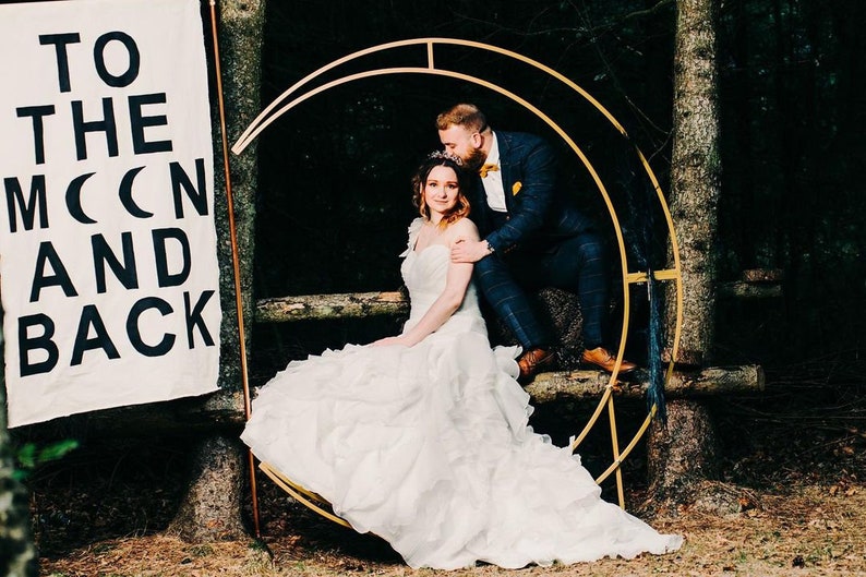 May include: A bride and groom pose for a wedding photo in a forest setting. The bride is wearing a white wedding dress and the groom is wearing a dark suit. They are standing in front of a gold metal archway decorated with greenery. The sign behind them reads "To the moon and back".