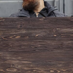 May include: A rectangular wooden sign with a dark brown finish. The wood grain is visible, and the sign is held by a person. The sign is made of several horizontal wooden planks.