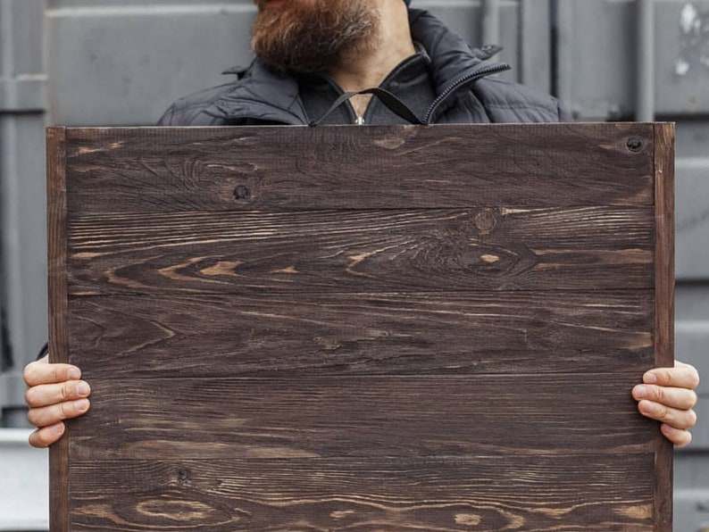 May include: A rectangular wooden sign with a dark brown finish. The wood grain is visible, and the sign is held by a person. The sign is made of several horizontal wooden planks.