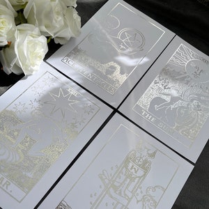 May include: Five tarot cards with silver foil designs on white card stock. The cards are: Ace of Pentacles, The Sun, Ace of Cups, The Star, and The World.