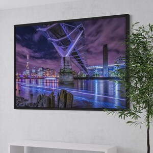 Puede incluir: Una fotografía enmarcada de un horizonte urbano de noche con un puente en primer plano. El puente está iluminado con luces moradas y azules, y el horizonte urbano se refleja en el agua debajo. La fotografía está colgada en una pared blanca en un interior moderno.
