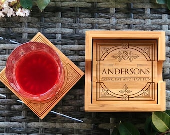 May include: A set of four bamboo coasters with a geometric design and the text "Andersons Drink, Eat and Have Fun". The coasters are on a woven surface with a glass of red liquid on one of the coasters.