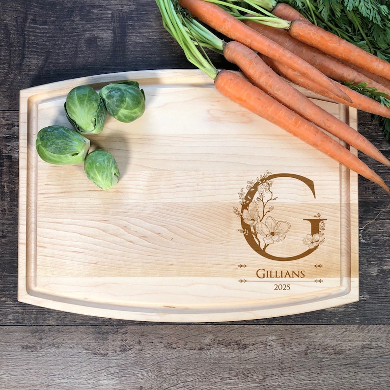 Puede incluir: Una tabla de cortar de madera con un borde curvo, que presenta un dise&ntilde;o floral con la letra "G" y el texto "Gillians 2025". La tabla se muestra con coles de Bruselas y zanahorias.