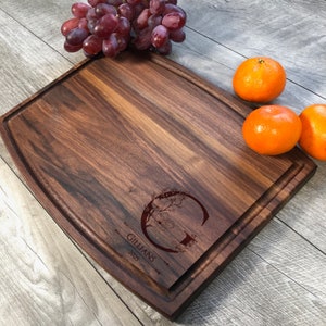 Puede incluir: Una tabla de cortar grande, rectangular, de madera de nogal con un borde redondeado. La tabla tiene un dise&ntilde;o grabado decorativo con la letra "G" y el nombre "Gillians" con el a&ntilde;o "2023" debajo. La tabla est&aacute; sobre una superficie de madera con naranjas y uvas a su lado.
