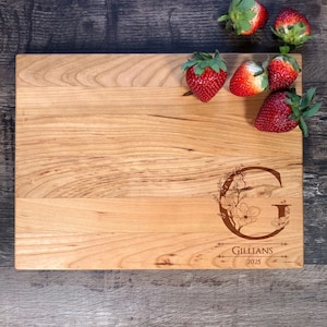 Puede incluir: Una tabla de cortar de madera personalizada con un dise&ntilde;o floral y la letra "G" grabada en ella. La tabla tambi&eacute;n tiene el nombre "Gillians" y el a&ntilde;o "2025" grabados en ella. La tabla est&aacute; rodeada de fresas.