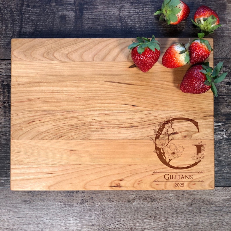 Puede incluir: Una tabla de cortar de madera personalizada con un dise&ntilde;o floral y la letra "G" grabada en ella. La tabla tambi&eacute;n tiene el nombre "Gillians" y el a&ntilde;o "2025" grabados en ella. La tabla est&aacute; rodeada de fresas.