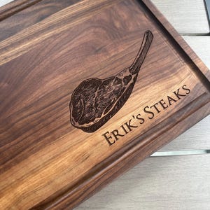 May include: A rectangular wooden chopping board with a carved illustration of a steak. The board is engraved with the text "Erik's Steaks". The wood has a rich, dark brown colour.