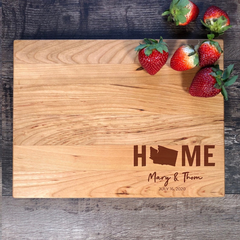 May include: A wooden chopping board with the words "HOME" and a silhouette of the state of Washington. The text "Mary & Thom" and "JULY 16, 2020" are also engraved on the board. There are strawberries on the surface of the board.