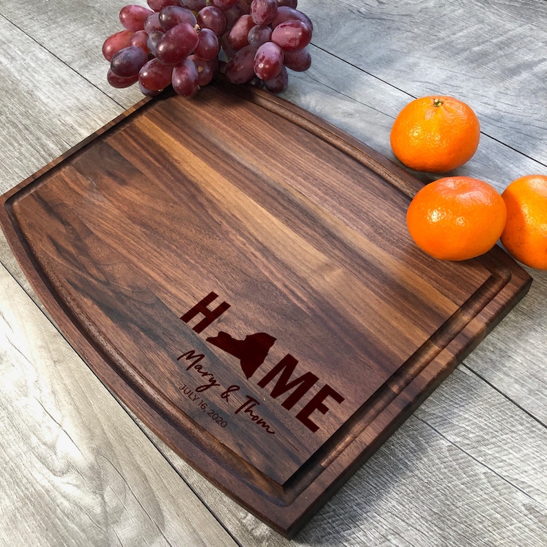 May include: A large, rectangular, wooden chopping board with a rounded edge. The board is made of walnut wood and has a natural finish. The board is engraved with the words "HOME" and "Mary & Thom" and the date "July 16, 2020". The board is also engraved with the outline of the state of New York. The board is sitting on a wooden surface with some fruit on the side.