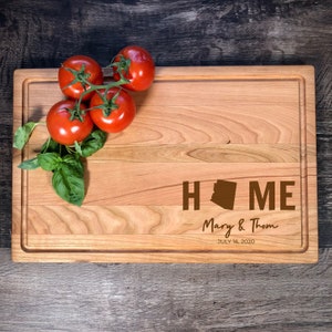 May include: A wooden chopping board with a brown outline and a brown "HOME" inscription with an Arizona state silhouette in the "O". The text "Mary & Thom JULY 16, 2020" is also engraved on the board. There are four red tomatoes on a vine in the top left corner of the board.
