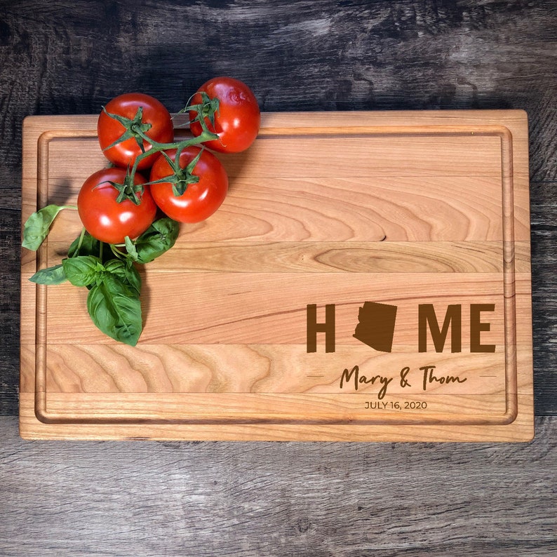 May include: A wooden chopping board with a brown outline and a brown "HOME" inscription with an Arizona state silhouette in the "O". The text "Mary & Thom JULY 16, 2020" is also engraved on the board. There are four red tomatoes on a vine in the top left corner of the board.