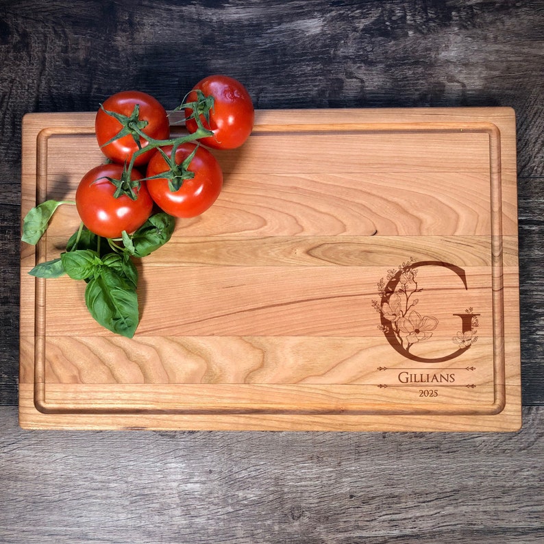 Puede incluir: Una tabla de cortar de madera personalizada con un dise&ntilde;o floral y la letra "G" grabada en ella. La tabla tambi&eacute;n tiene el nombre "Gillians" y el a&ntilde;o "2023" grabados en ella. La tabla est&aacute; hecha de madera de cerezo y tiene un acabado natural.