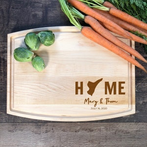 May include: A wooden chopping board with a brown "HOME" inscription, featuring a state outline, and the text "Mary & Thom JULY 16, 2020". The board has a rounded edge and is on a wooden surface with green brussels sprouts.