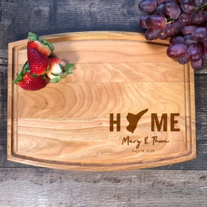 May include: A wooden chopping board with a curved edge, engraved with the word "HOME" and a state outline. The text "Mary & Thom JULY 16, 2020" is also engraved on the board. There are strawberries on the board.