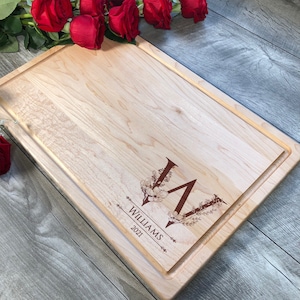 Puede incluir: Una tabla de cortar de madera de arce personalizada con un dise&ntilde;o de monograma floral. El monograma es la letra "V" con un dise&ntilde;o floral y el texto "Williams 2021".