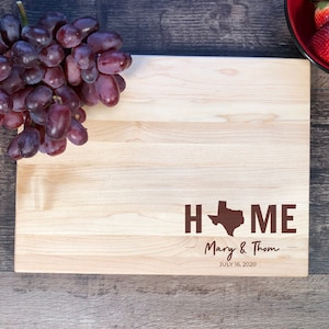 May include: A light brown wooden chopping board with the words "HOME" in brown text, with a silhouette of the state of Texas in the middle of the word "HOME". The text "Mary & Thom" and "JULY 16, 2020" is also engraved on the board.