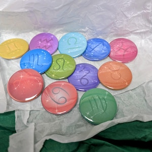Puede incluir: Una colección de diez botones redondos y coloridos con signos del zodiaco. Cada botón presenta un símbolo astrológico diferente en un color contrastante, sobre un fondo degradado. Los botones están dispuestos sobre papel de seda blanco, con un fondo verde.