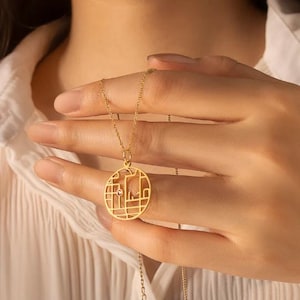 Puede incluir: Un collar de tono dorado con un colgante circular que presenta un intrincado patrón de rejilla calada similar a un mapa. El colgante incluye una pequeña piedra preciosa redonda de color rosa engastada en la estructura metálica, suspendido de una delicada cadena dorada.