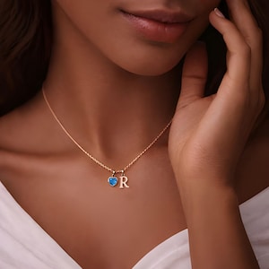 Puede incluir: Un collar de cadena en tono dorado con dos pequeños dijes: un colgante en forma de corazón azul brillante y un colgante con la letra mayúscula R. El collar descansa sobre el cuello, mostrando un diseño de cadena fina con acabados de metal pulido.
