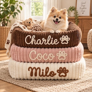 Puede incluir: Una pila de tres camas rectangulares para mascotas en tela de pana marrón, rosa y blanca. Cada cama presenta una textura de felpa, un icono de huella bordado y un nombre en la parte frontal: Charlie, Coco y Milo. Un perro pequeño está sentado encima.