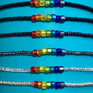May include: Seven rainbow colored beaded bracelets with different colored strings. The bracelets are arranged in a vertical stack, with the top bracelet being black and the bottom bracelet being white.