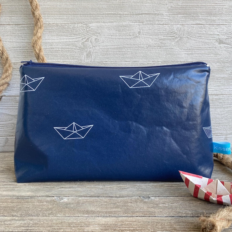 Könnte beinhalten: Eine dunkelblaue Reißverschlusstasche mit einem weißen Papierboot-Design. Die Tasche ist aus einem strukturierten Material gefertigt und hat einen weißen Reißverschluss.