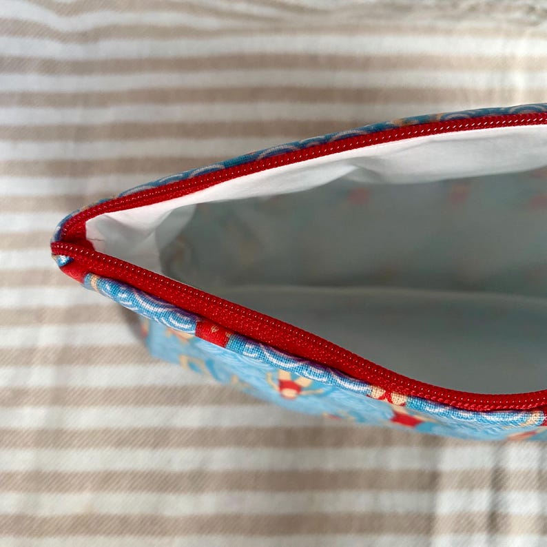Peut inclure: Une pochette rectangulaire partiellement ouverte avec une fermeture &eacute;clair rouge et un ext&eacute;rieur bleu clair. L'ext&eacute;rieur de la pochette pr&eacute;sente un motif de petites formes color&eacute;es. L'int&eacute;rieur est blanc et la fermeture &eacute;clair est rouge.