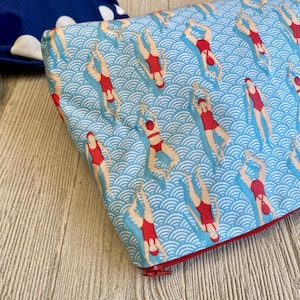 Peut inclure: Une pochette rectangulaire avec une fermeture &eacute;clair rouge, orn&eacute;e d'un motif r&eacute;p&eacute;t&eacute; de nageurs en maillots de bain rouges sur un fond bleu &agrave; vagues. La pochette est probablement en tissu.