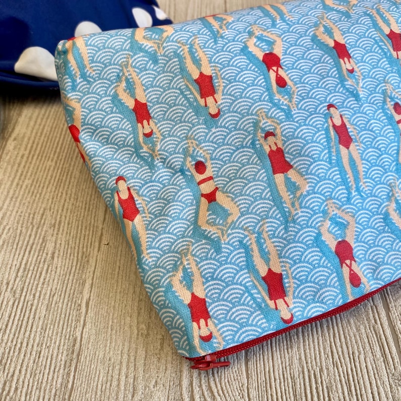 Peut inclure: Une pochette rectangulaire avec une fermeture &eacute;clair rouge, orn&eacute;e d'un motif r&eacute;p&eacute;t&eacute; de nageurs en maillots de bain rouges sur un fond bleu &agrave; vagues. La pochette est probablement en tissu.