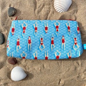 Könnte beinhalten: Eine blaue und weiße Stofftasche mit einem roten und weißen Muster von Schwimmern. Die Tasche hat einen roten Reißverschluss und liegt auf einer sandigen Oberfläche.