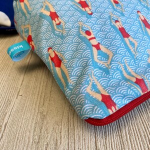 Peut inclure: Une pochette bleue &agrave; fermeture &eacute;clair avec un motif r&eacute;p&eacute;t&eacute; de nageurs en maillots de bain rouges sur un fond bleu ondul&eacute;. La pochette a une fermeture &eacute;clair rouge et une petite &eacute;tiquette bleue avec le mot "Kiss".