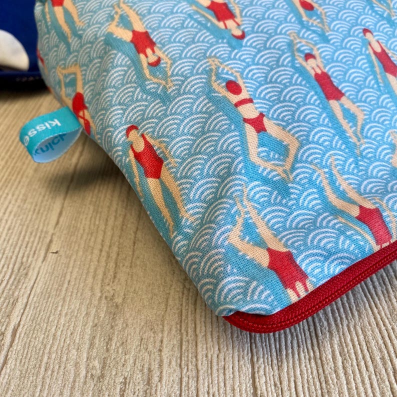 Peut inclure: Une pochette bleue &agrave; fermeture &eacute;clair avec un motif r&eacute;p&eacute;t&eacute; de nageurs en maillots de bain rouges sur un fond bleu ondul&eacute;. La pochette a une fermeture &eacute;clair rouge et une petite &eacute;tiquette bleue avec le mot "Kiss".