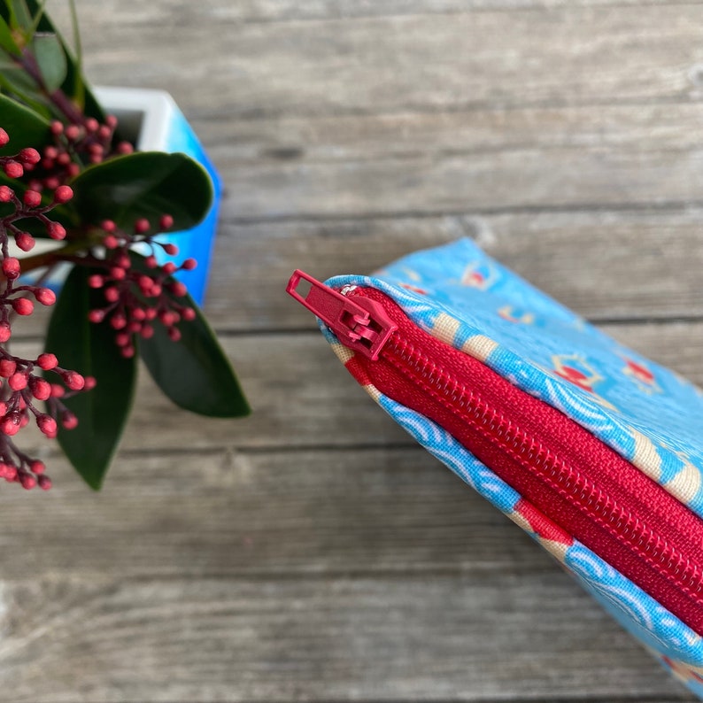 Peut inclure: Gros plan sur une pochette en tissu bleu avec une fermeture &eacute;clair rouge. La pochette pr&eacute;sente un motif r&eacute;p&eacute;titif de formes rouges et jaunes sur un fond bleu clair. La fermeture &eacute;clair est ouverte, r&eacute;v&eacute;lant l'int&eacute;rieur rouge.