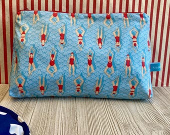 Peut inclure: Une pochette rectangulaire bleu clair avec fermeture éclair, ornée d'un motif répété de nageuses synchronisées en maillots de bain rouges. Les nageuses sont disposées sur un fond d'eau ondulée. La pochette porte une petite étiquette bleue avec le mot "kiss".