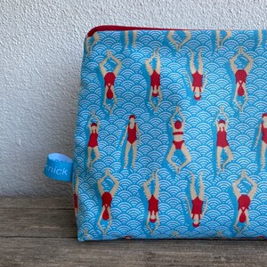 Peut inclure: Une trousse de maquillage rectangulaire avec un motif de vagues bleues et des nageurs en rouge. La trousse a une fermeture &eacute;clair rouge et une petite &eacute;tiquette bleue avec le mot "knick".