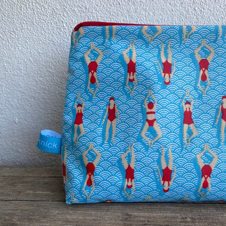 Peut inclure: Une trousse de maquillage rectangulaire avec un motif de vagues bleues et des nageurs en rouge. La trousse a une fermeture &eacute;clair rouge et une petite &eacute;tiquette bleue avec le mot "knick".