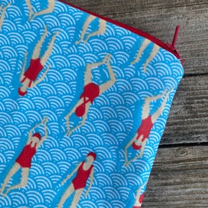 Peut inclure: Une pochette bleue &agrave; fermeture &eacute;clair avec un motif de nageurs en maillots de bain rouges plongeant dans l'eau. L'eau est repr&eacute;sent&eacute;e par des motifs de vagues blanches sur fond bleu. La fermeture &eacute;clair est bordeaux.