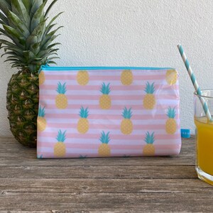 Könnte beinhalten: Eine rosa-weiße gestreifte Reißverschlusstasche mit einem sich wiederholenden Muster aus gelben Ananas. Die Tasche ist aus einem klaren, glänzenden Material.