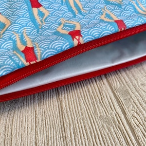 Peut inclure: Une pochette zipp&eacute;e avec un fond bleu orn&eacute; d'un motif de nageuses synchronis&eacute;es en maillots de bain rouges. La pochette a une fermeture &eacute;clair et une bordure rouges, et un int&eacute;rieur gris clair. La pochette est sur une surface textur&eacute;e beige clair.
