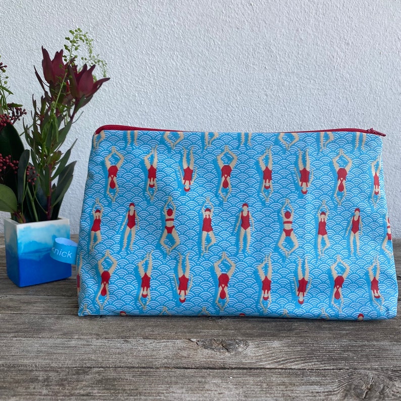 Peut inclure: Une trousse de maquillage rectangulaire avec un fond bleu orn&eacute; d'un motif de nageurs en maillots de bain rouges. Le sac a une fermeture &eacute;clair rouge et un motif de vagues bleues et blanches. Le sac est sur une surface en bois.