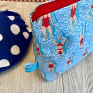 Peut inclure: Une trousse de maquillage bleue avec une fermeture &eacute;clair rouge et un motif de nageurs en maillots de bain rouges. Le sac a une &eacute;tiquette bleue avec le mot "knick". Un sac bleu et blanc &agrave; pois est visible en arri&egrave;re-plan.