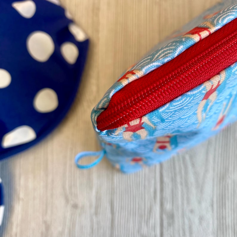 Peut inclure: Une trousse de maquillage bleue avec une fermeture &eacute;clair rouge et une petite boucle. Le sac pr&eacute;sente un motif de nageurs et de vagues. Un article bleu &agrave; pois blancs est visible en arri&egrave;re-plan.